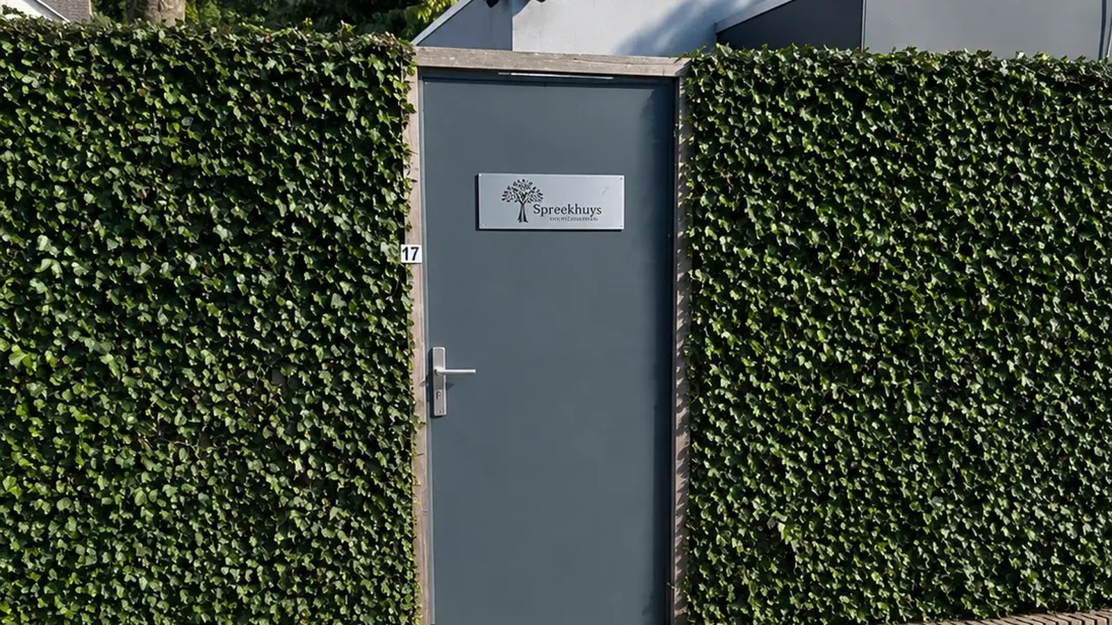 De entree van Spreekhuys in Boxmeer: grijze deur met naambord tussen groene hagen